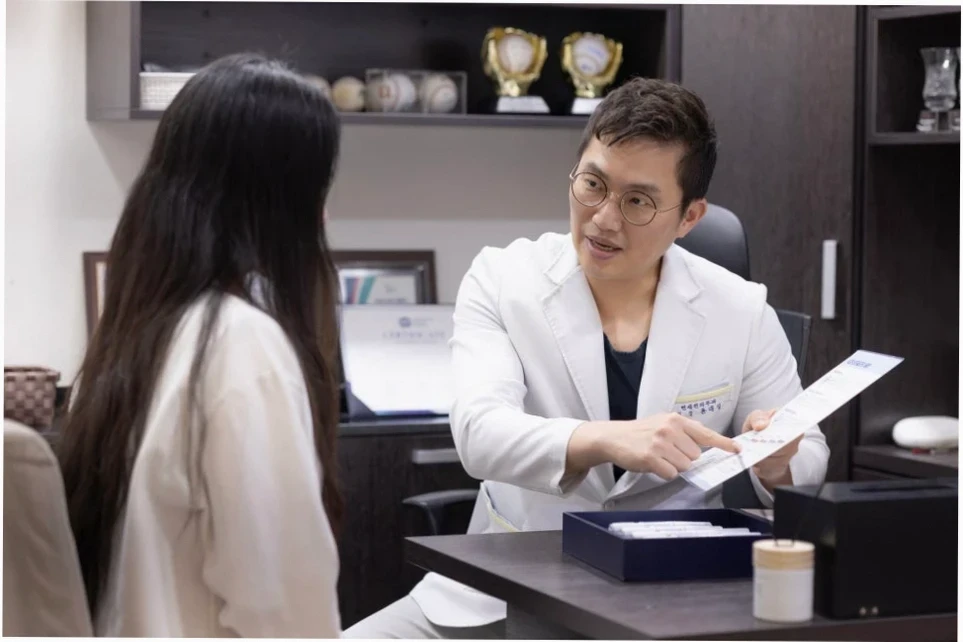 흰 가운을 입은 남성 의사가 안경을 쓴 채 여성 환자와 앉아 상담하며 서류를 가리키고 있습니다. 신뢰할 수 있는 목동 피부과 상담 분위기를 묘사합니다.