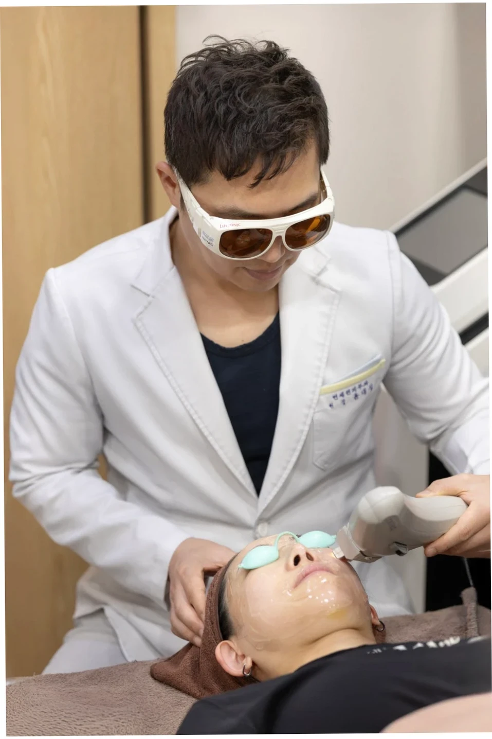 보호 안경을 쓴 의사가 누워있는 환자의 얼굴에 젤을 바르고 레이저 시술을 하는 장면입니다. 전문 장비로 피부 고민을 해결하는 목동 피부과의 모습을 보여줍니다.